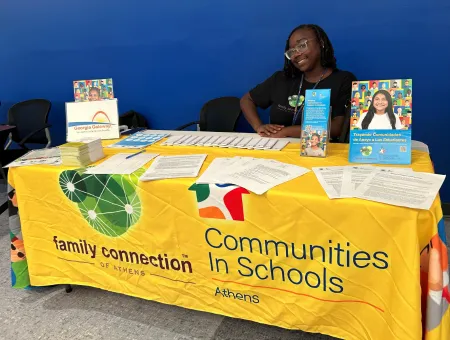 Zowee sitting at a table with a yellow cloth at new student orientation.